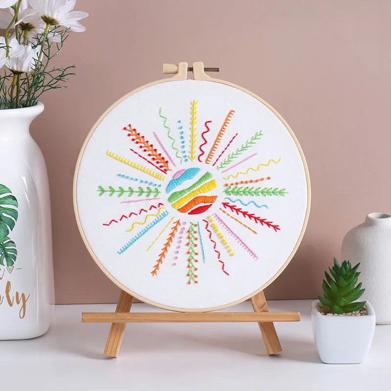 Embroidery hoop with colorful patterns on a wooden stand against a pink wall.