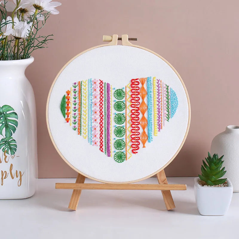 Embroidery hoop with colorful patterns on a stand against a pink wall with decorative items.