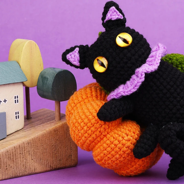 Crocheted black cat with yellow eyes holding an orange pumpkin against a purple background