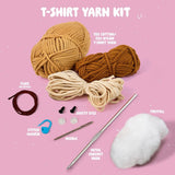 T-shirt yarn kit with yarn balls, safety eyes, needle, metal crochet hook, and polyfill on a pink background.