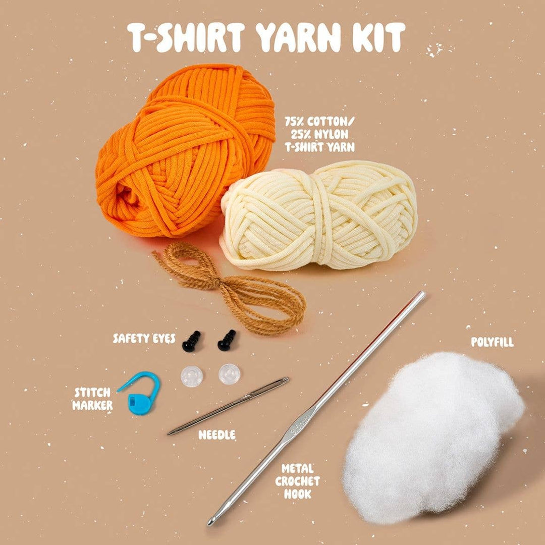 T-shirt yarn kit with orange and white yarn, safety eyes, polyfill, needle, metal crochet hook, and stitch marker on a beige background.