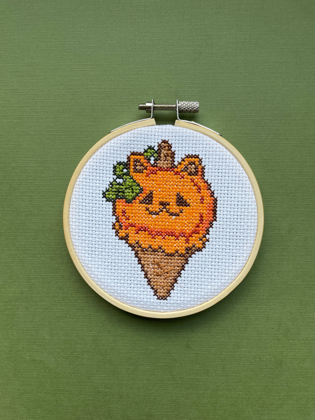 Cross-stitched design of a carrot with a face in an embroidery hoop on a green background