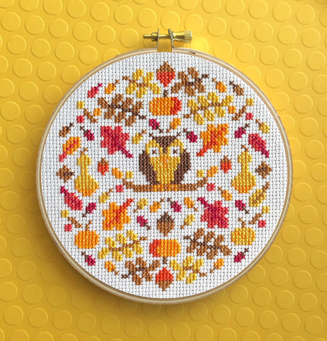 Cross-stitched pattern with an owl and autumn leaves in a hoop on a yellow background