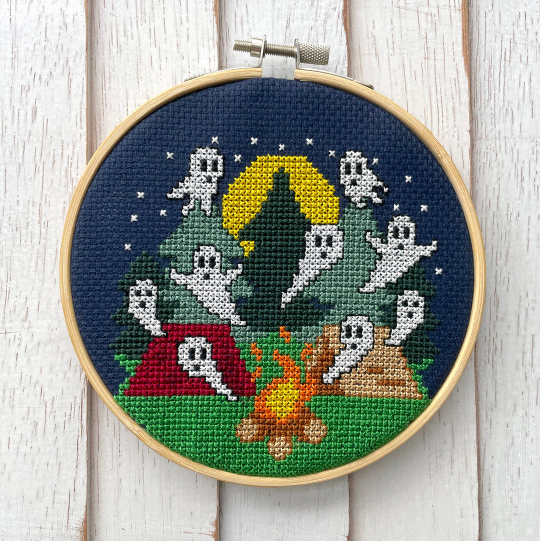 Embroidery hoop with a Halloween-themed cross-stitch design of ghosts around a campfire.
