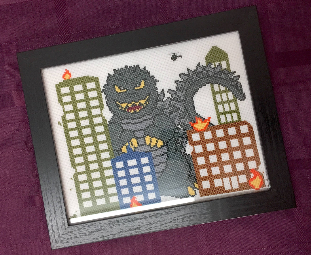 Framed cross-stitched picture of Godzilla destroying buildings on a purple background