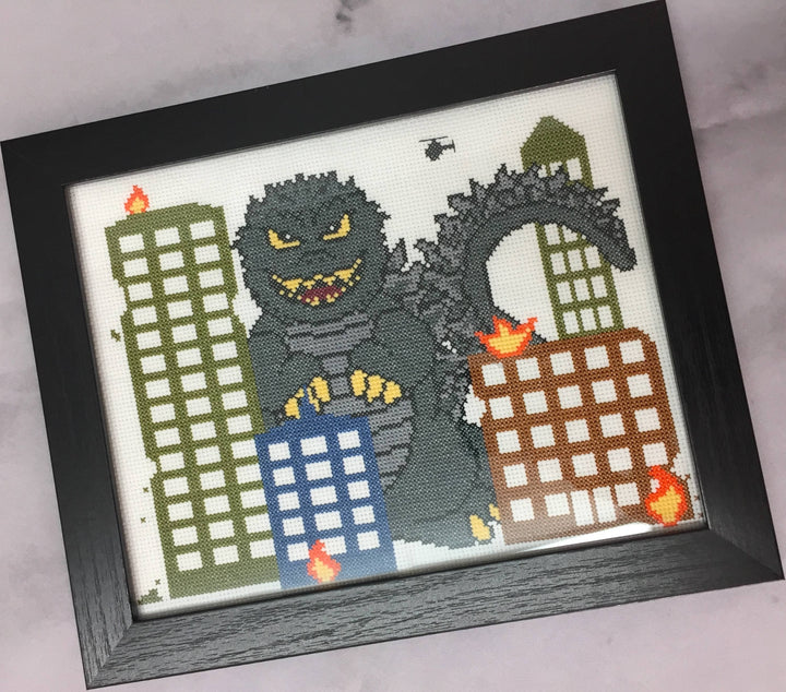 Framed cross-stitched picture of Godzilla destroying buildings