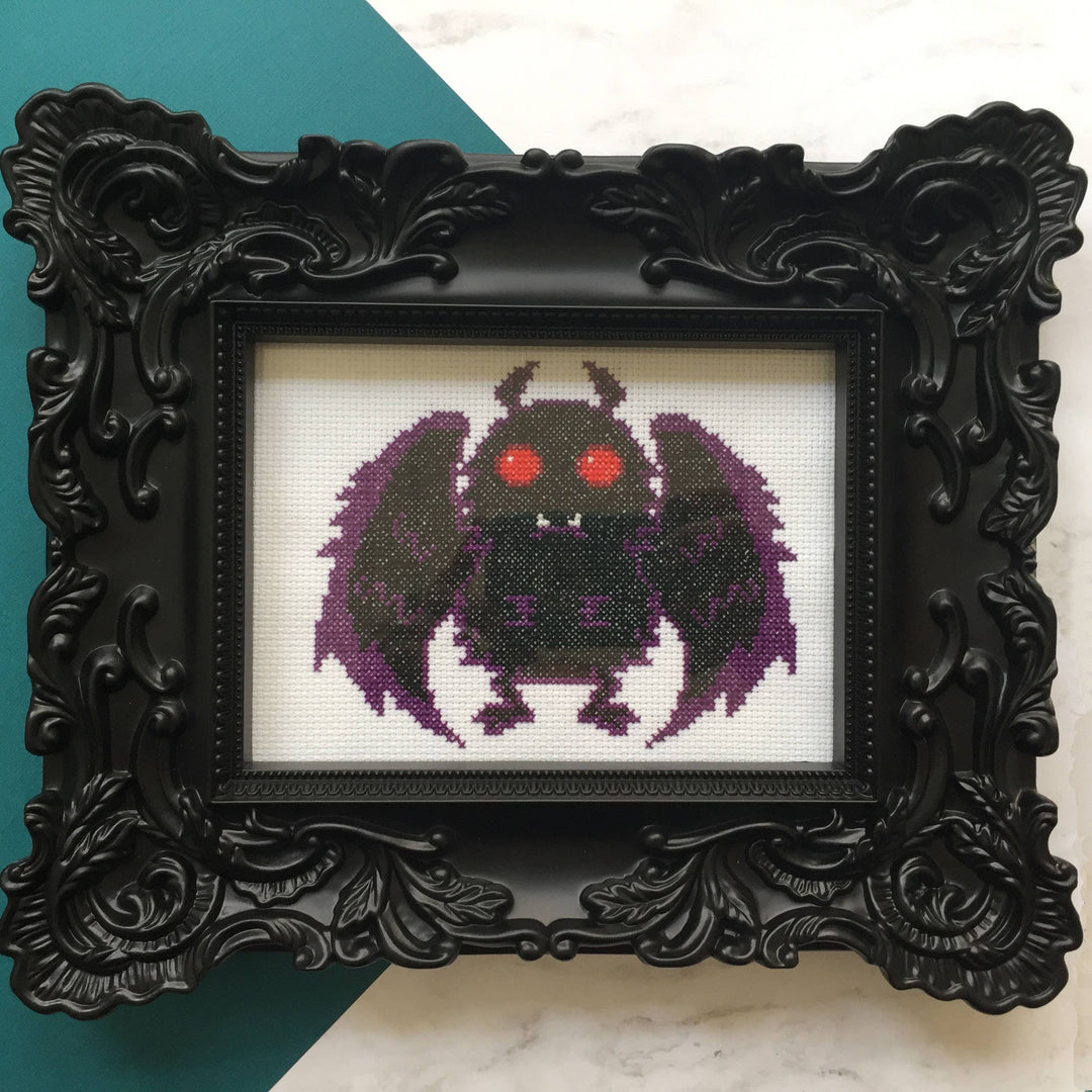 Framed cross-stitched purple creature with red eyes on a white background
