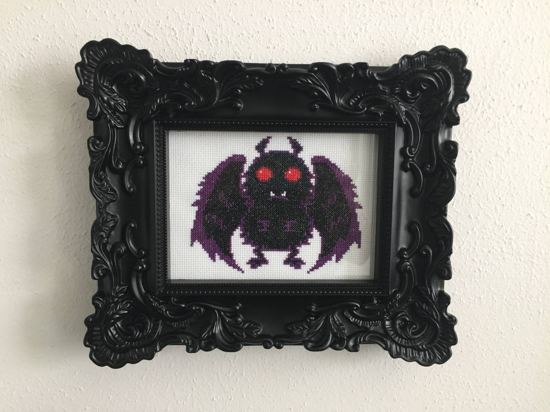 Framed cross-stitched artwork of a purple creature with red eyes on a white background