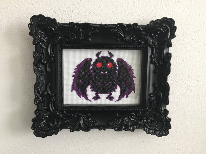 Framed cross-stitched artwork of a purple creature with red eyes on a white background
