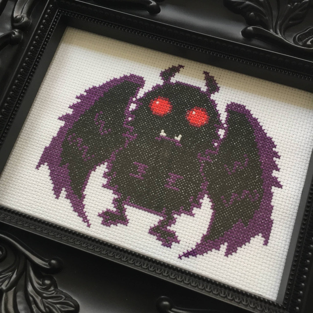 Cross-stitched creature with purple wings and red eyes on a white background, framed in black.