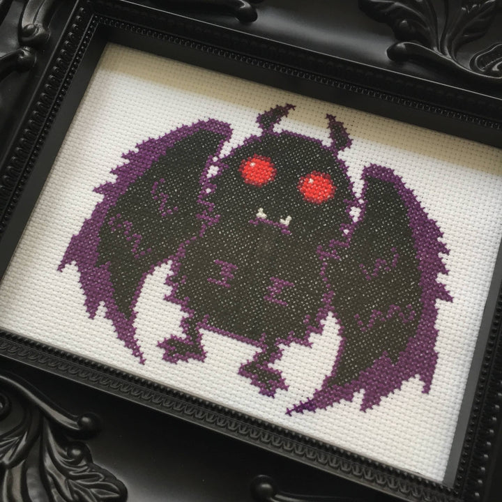 Cross-stitched creature with purple wings and red eyes on a white background, framed in black.