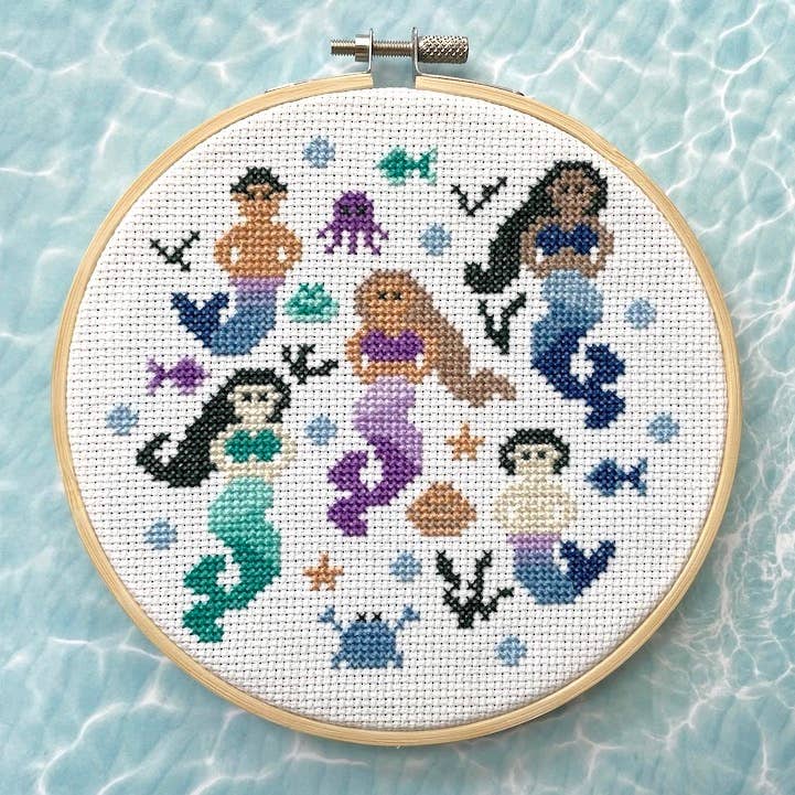 Cross-stitched design of mermaids and sea creatures in an embroidery hoop on a light blue water pattern background.
