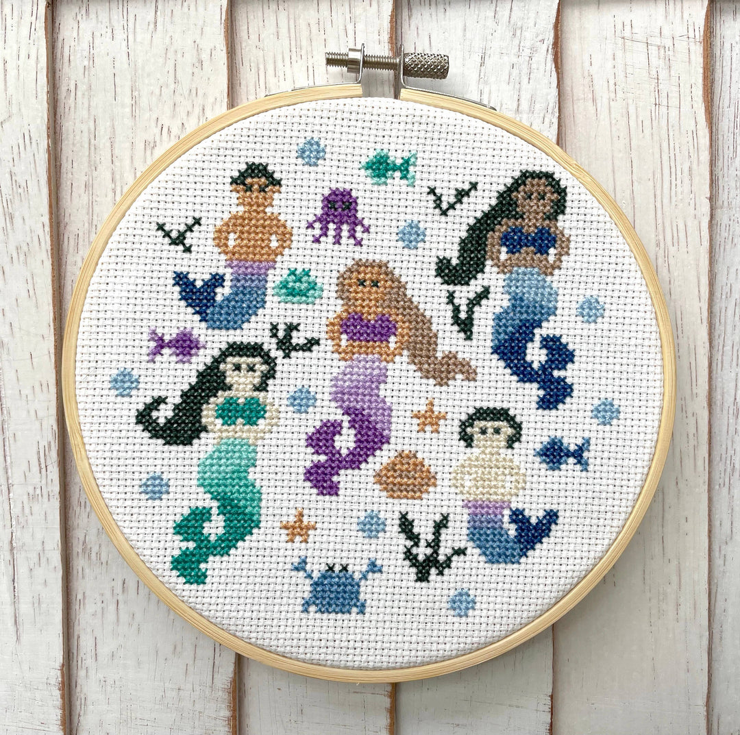 Cross-stitched design of mermaids and sea creatures in a wooden hoop on a wooden background