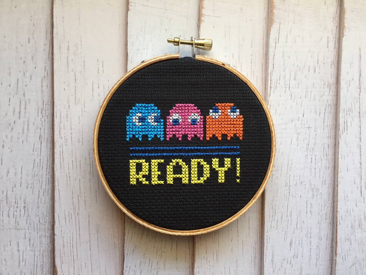 Cross-stitched design of Pac-Man ghosts and 'READY!' text in a wooden hoop on a light wood background.