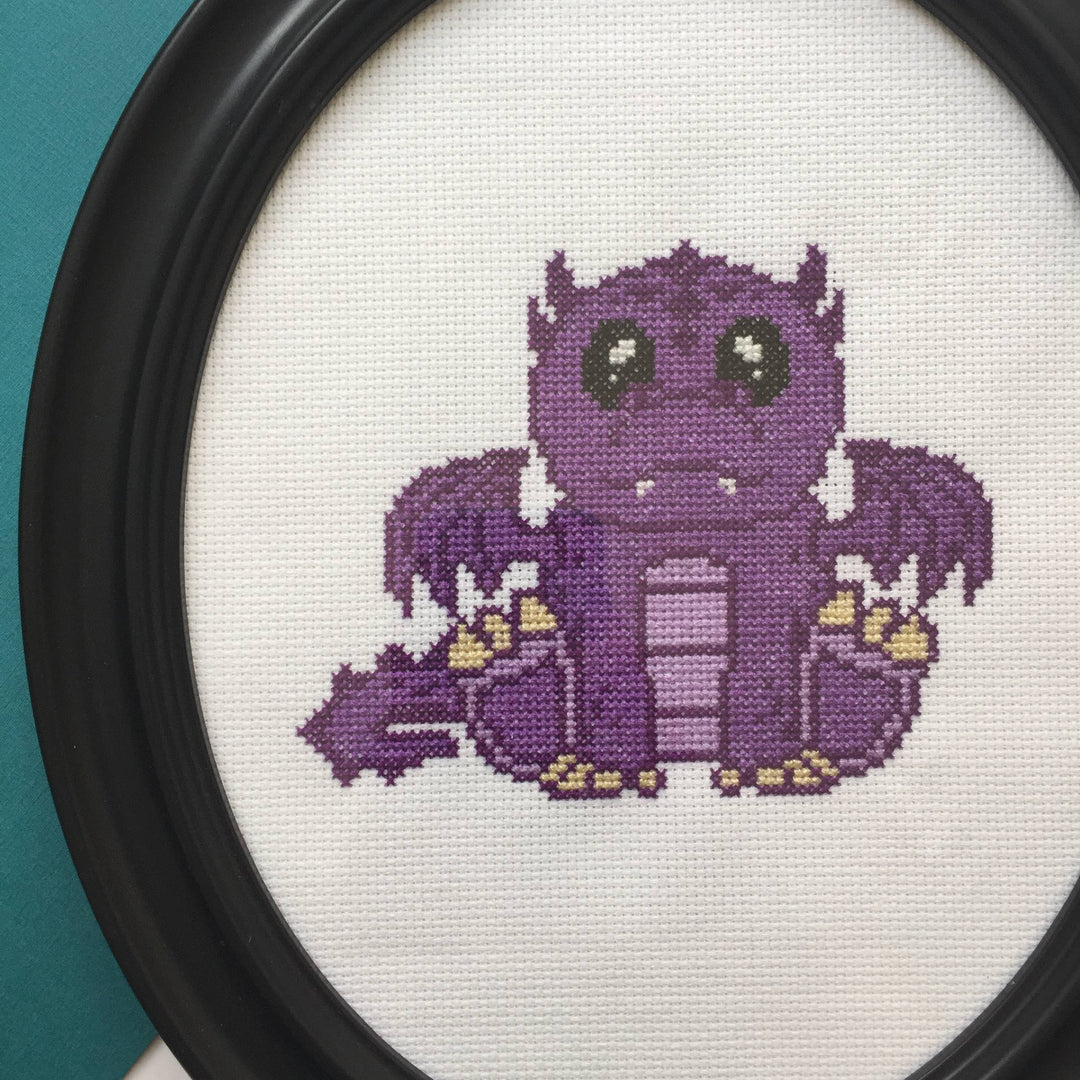 Cross-stitched purple creature with yellow accents in a black frame against a teal background
