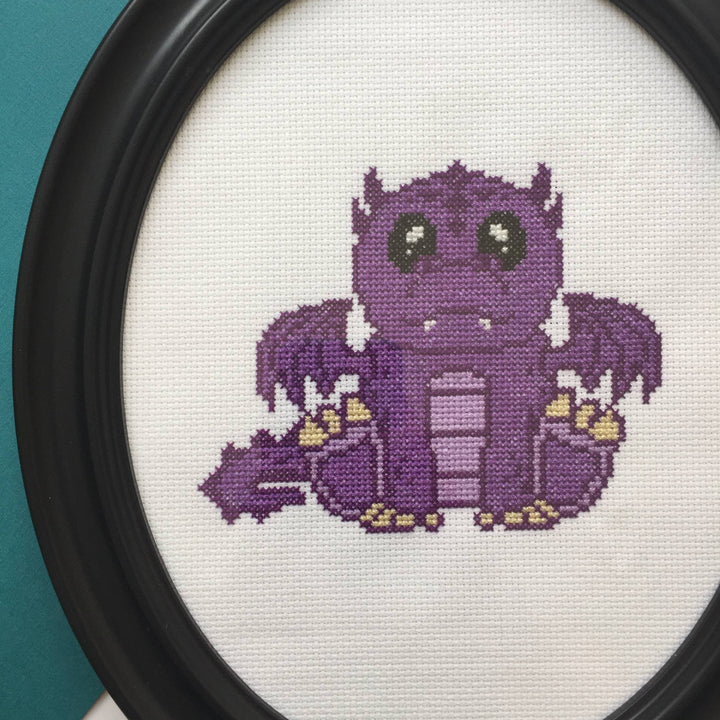 Cross-stitched purple creature with yellow accents in a black frame against a teal background