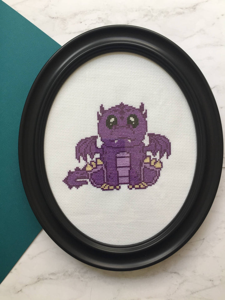 Framed cross-stitched purple creature on a white background