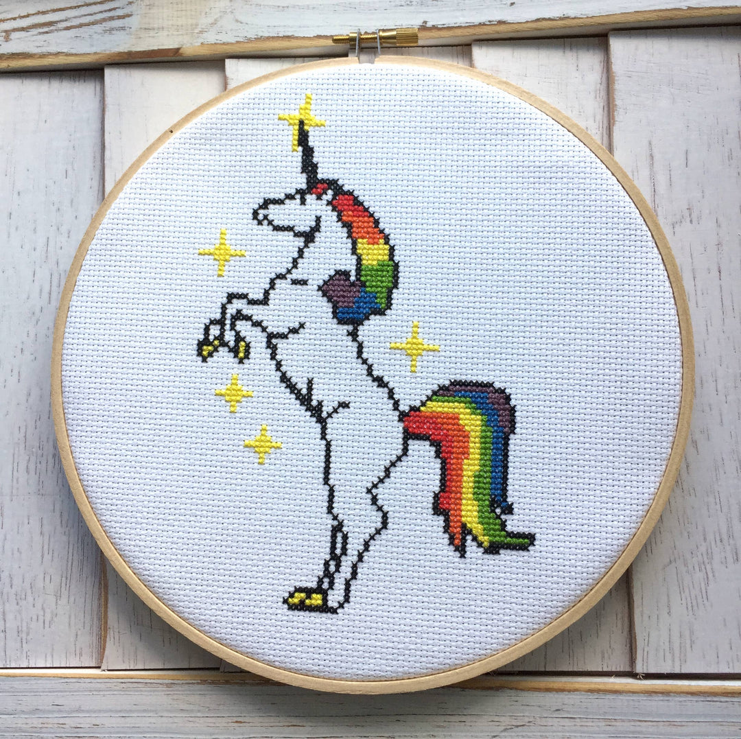 Cross-stitched unicorn with rainbow tail and horn in a wooden hoop on a light wooden background