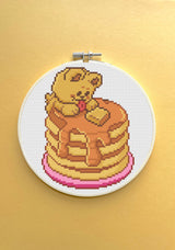 Cross-stitched design of a bear on pancakes with syrup in an embroidery hoop against a yellow background