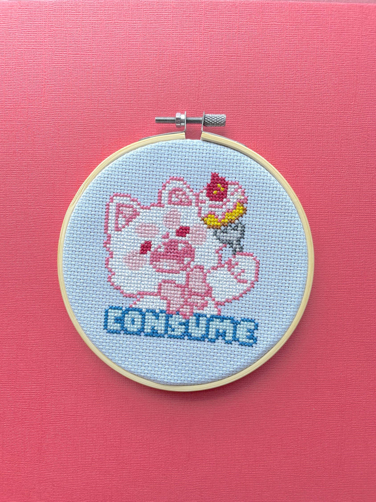 Cross-stitched design of a cartoon pig with a bow tie in an embroidery hoop on a pink background.