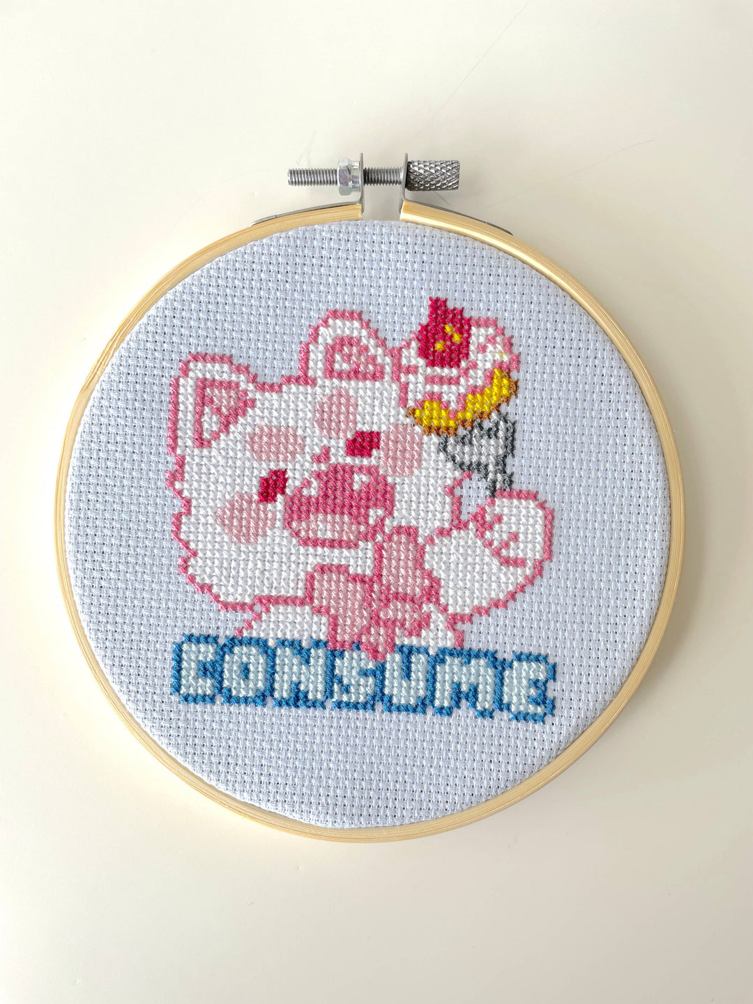 Cross-stitched design of a cartoon character with a cake on its head in an embroidery hoop.