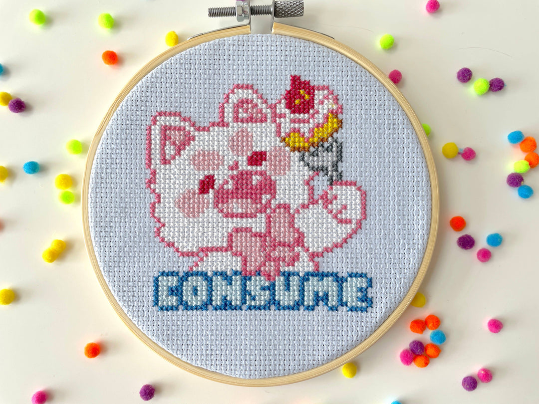 Cross-stitched design of a cartoon character with 'CONSUME' text in an embroidery hoop, surrounded by colorful beads.