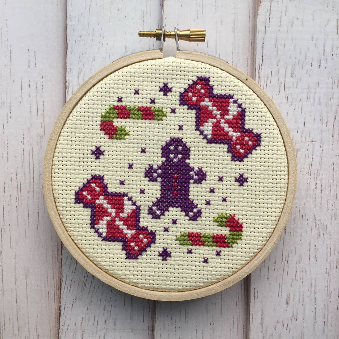 Cross-stitched gingerbread man and candy design in a wooden hoop on a light wooden surface.